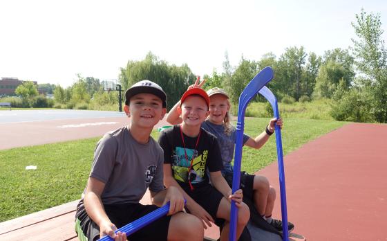 Three boys siting on a bench holding plastic hockey sticks smiling at the camera