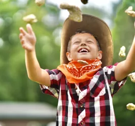 Kid smiling in a checked shirt, bandana, and cowboy hat throwing up "gold nuggets" laughing