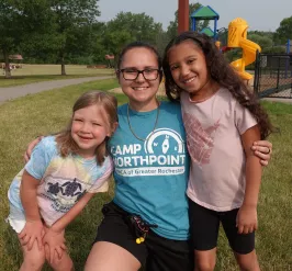 a camp northpoint staff member kneeling with two campers in the grass outside of the aquatic center, smiling at the camera