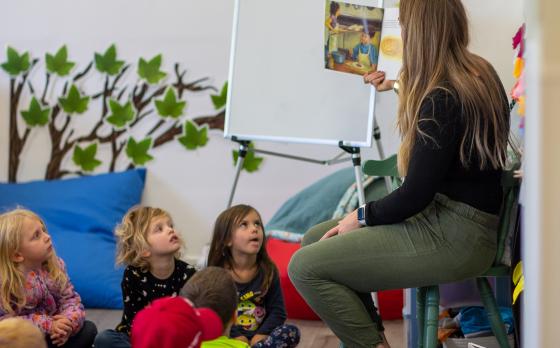 a teacher seated and reading a book to preschoolers sitting on the floor in front of her