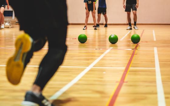 a photo of three green dodgeballs lined up on a gym floor with peoples legs running towards them
