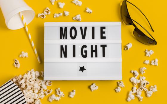 Yellow background with a frame reading "movie night" in the middle, surrounded by an empty cup, black sunglasses, black and white stripped popcorn containers and spilled popcorn