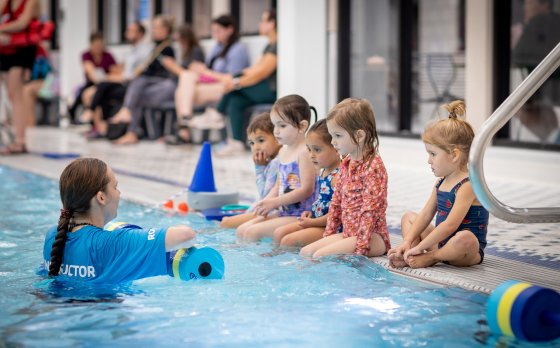 a swim instructor is in the pool speaking with a group of five young swimmers during a swim lesson