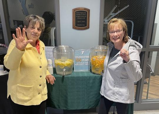 two women waving at the camera in front of drink dispensers filled with lemon and orange slices