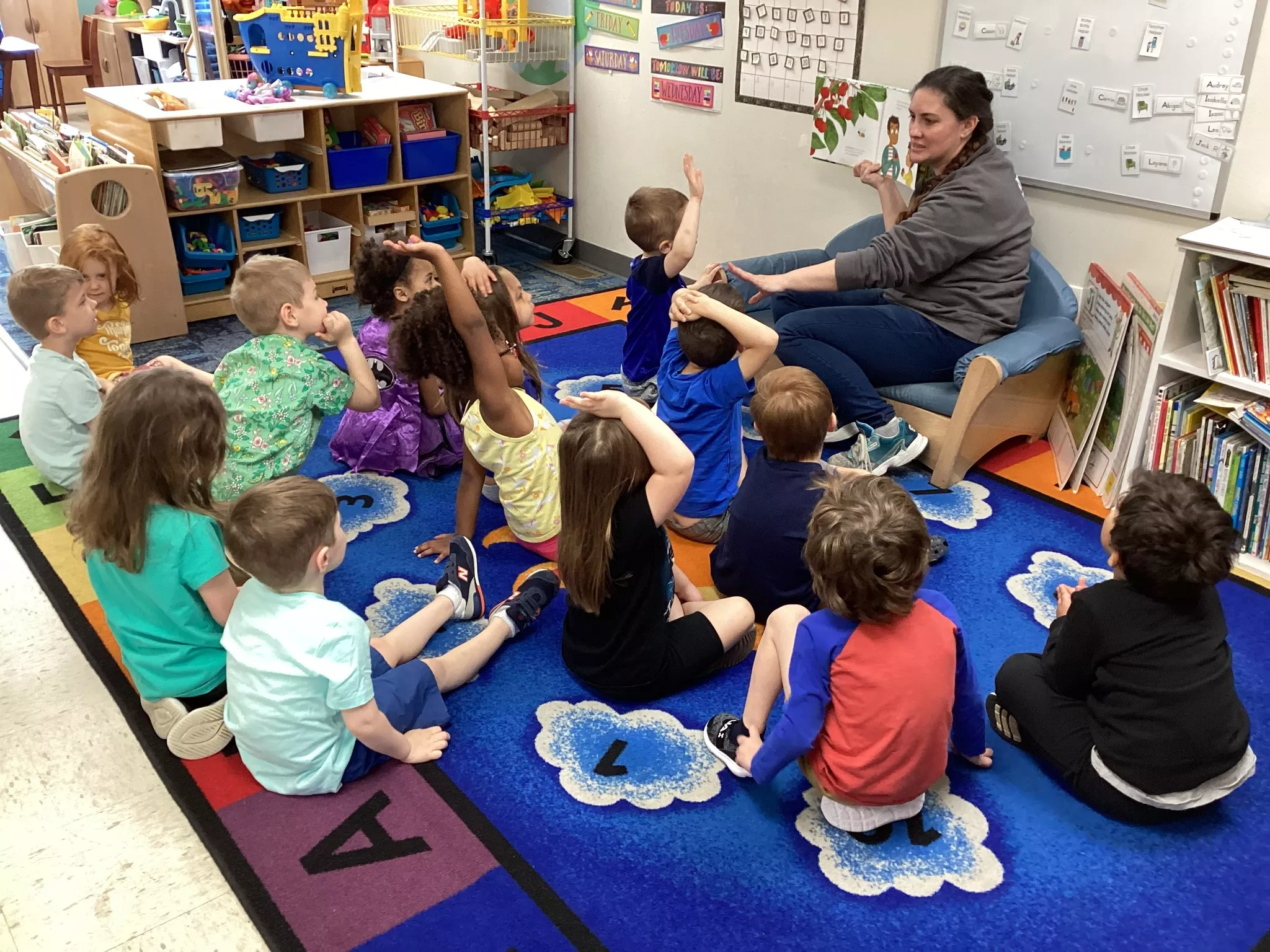 14 students sitting on a rug in front of a teacher reading them a book