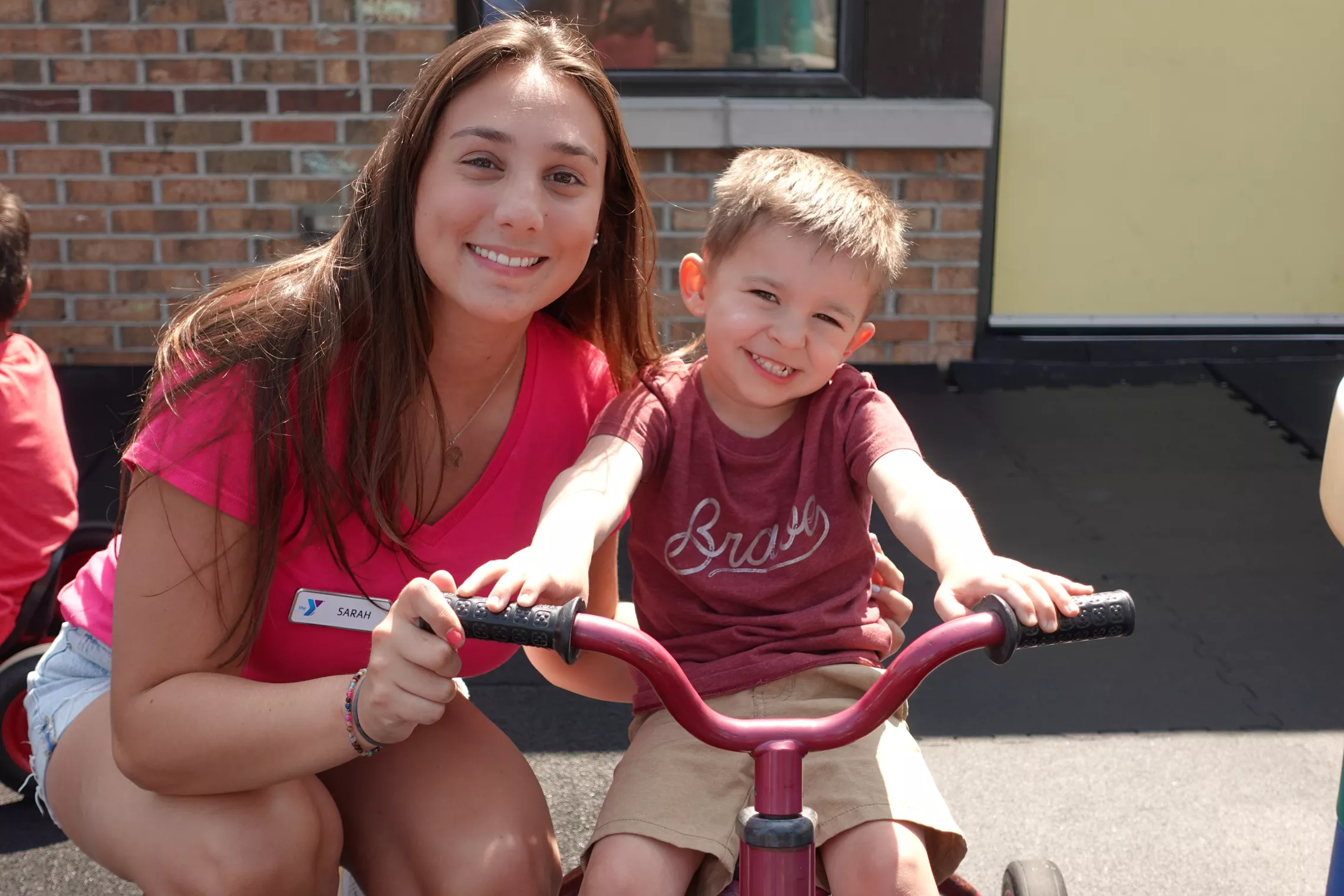 a young child sitting on a red tricycle outside with a young ymca staff member kneeling next to him.