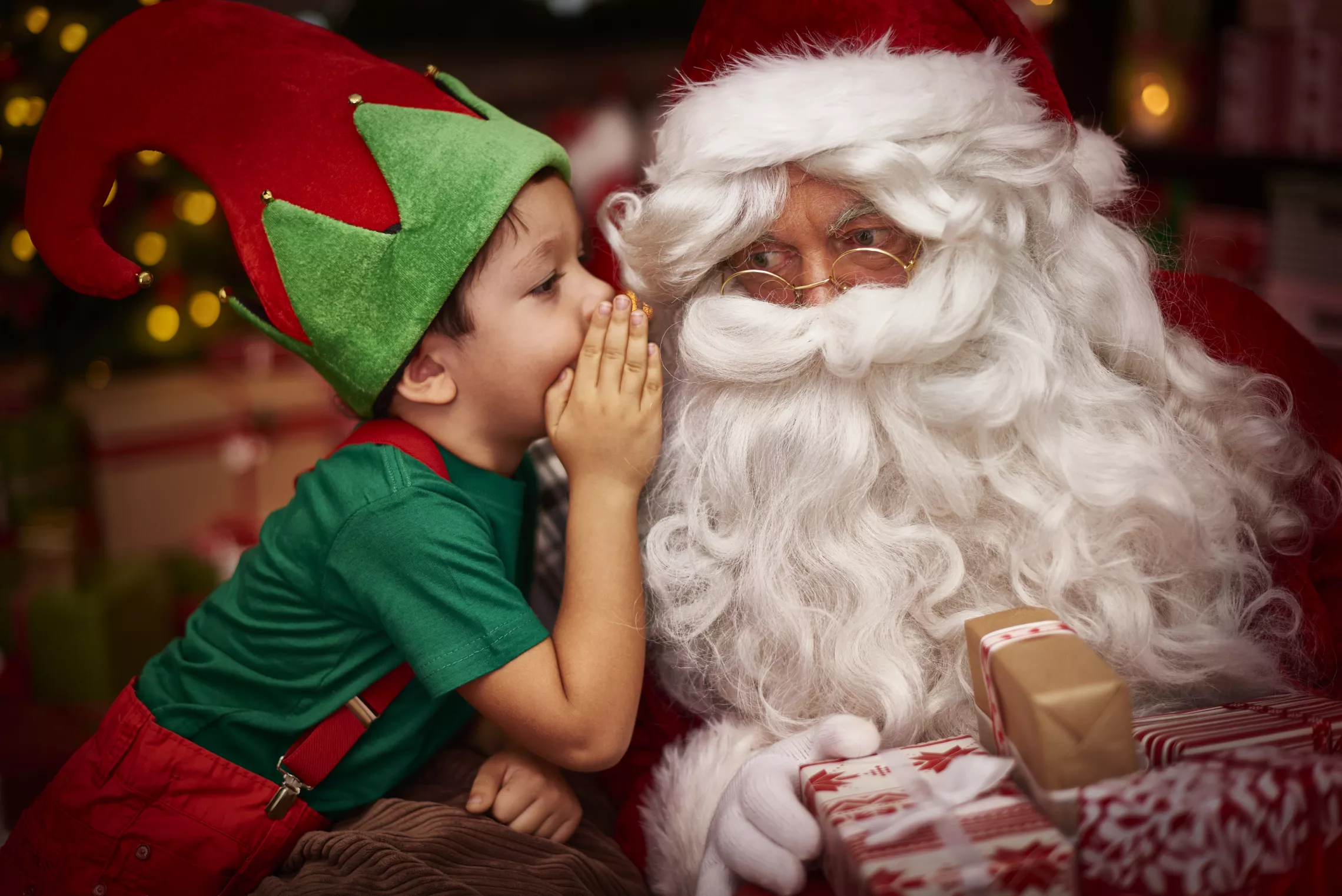 child in an elf costume leaning in and whispering in santa's ear while santa holds presents