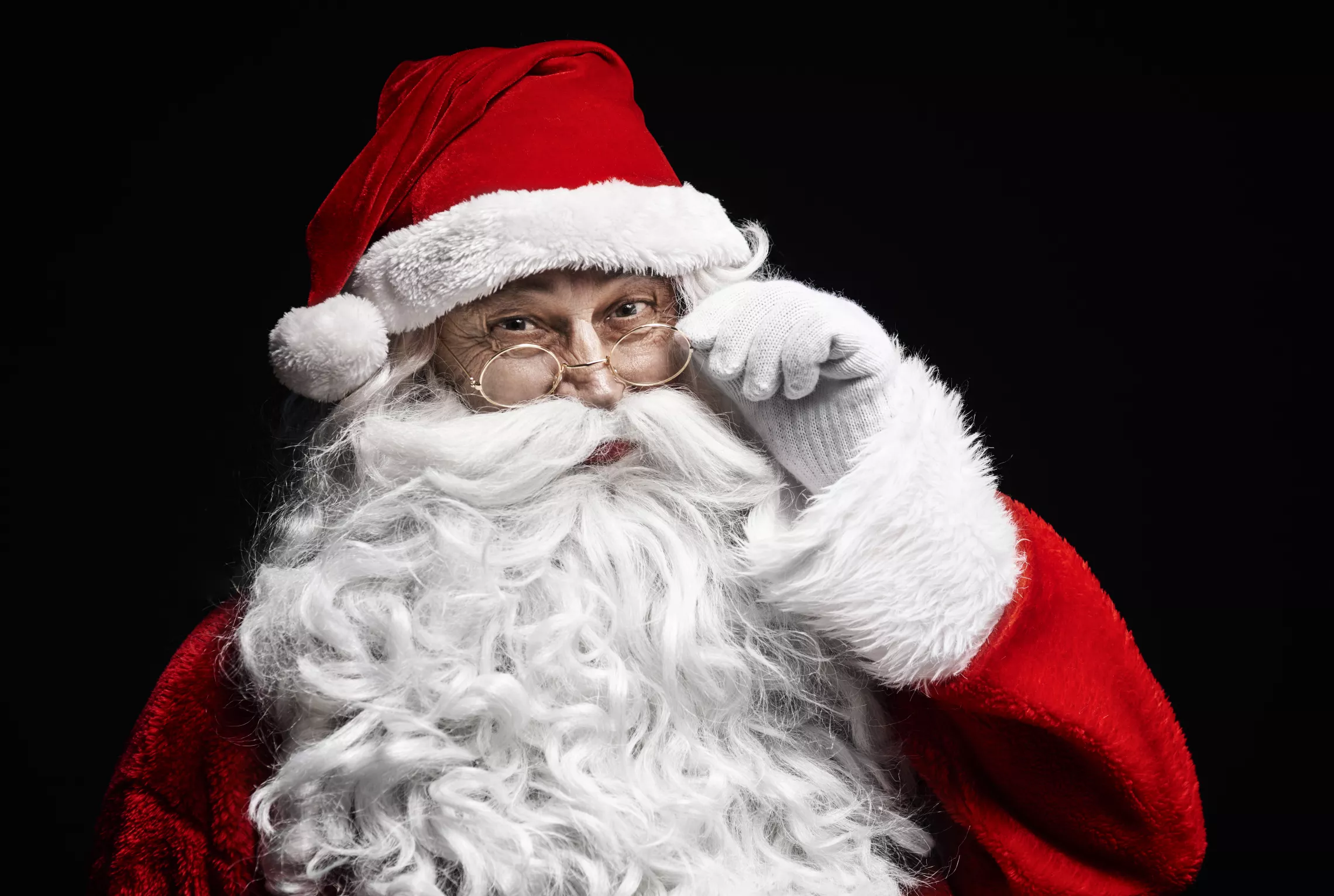 Santa wearing a read jacket with white cuffs and wire-rimmed glasses in front of a black background
