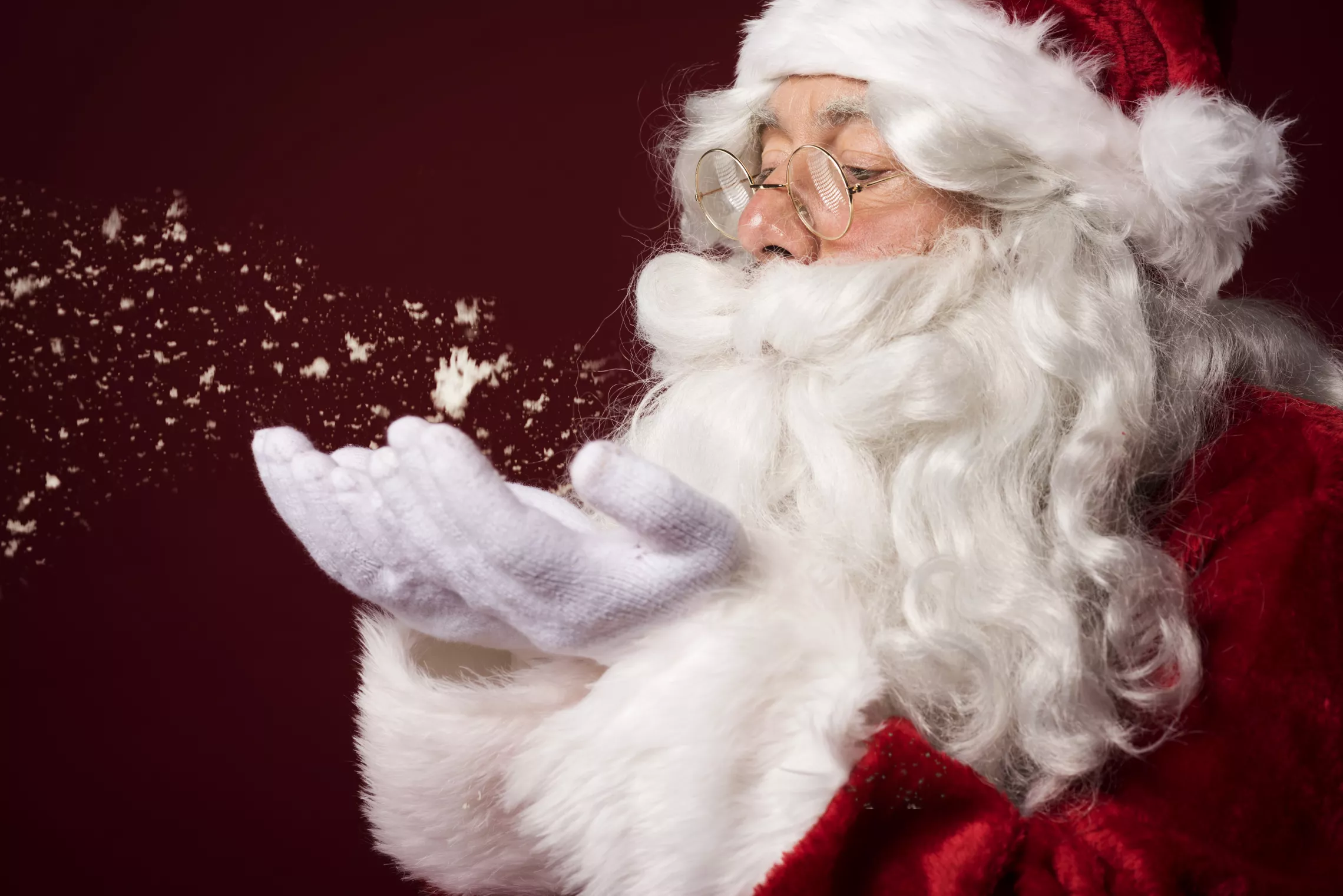 Santa Claus blowing snowflakes off of his hand in front of a red background