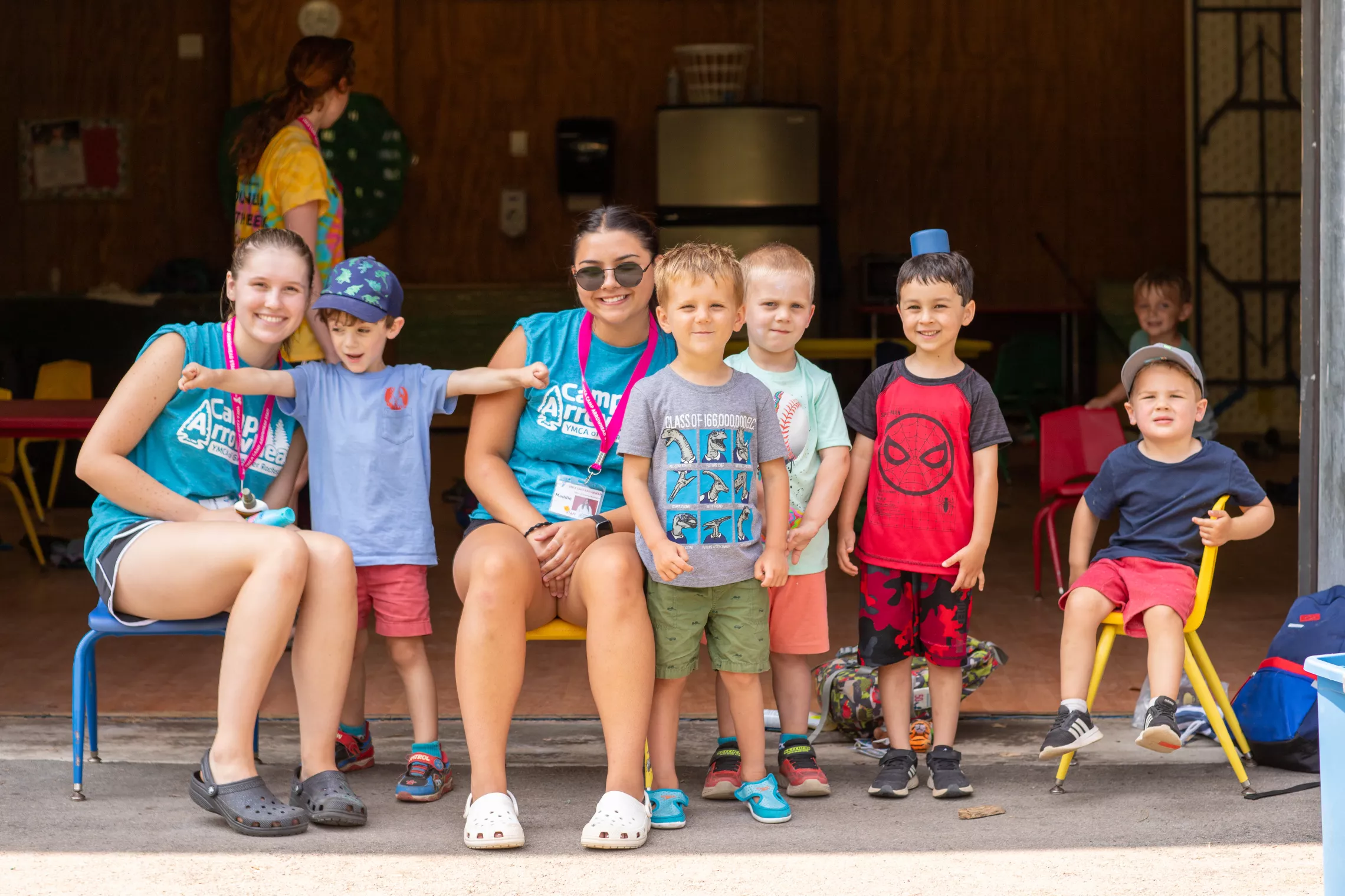 Two staff members witting with 5 preschool campers outside of the Oneida shelter smiling at the camera
