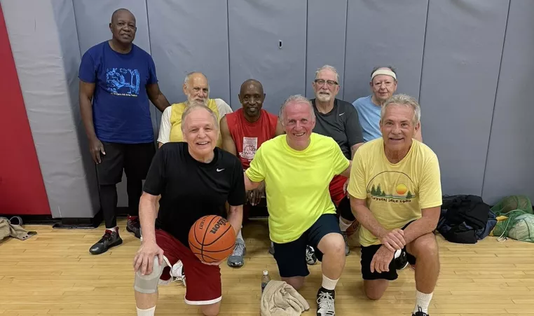 8 older men posing for a photo in the northwest gymnasium with a basketball