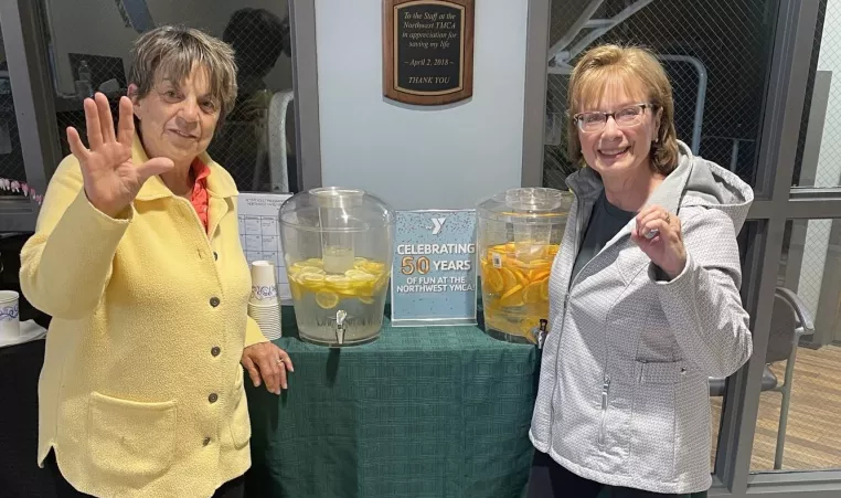 two women waving at the camera in front of drink dispensers filled with lemon and orange slices