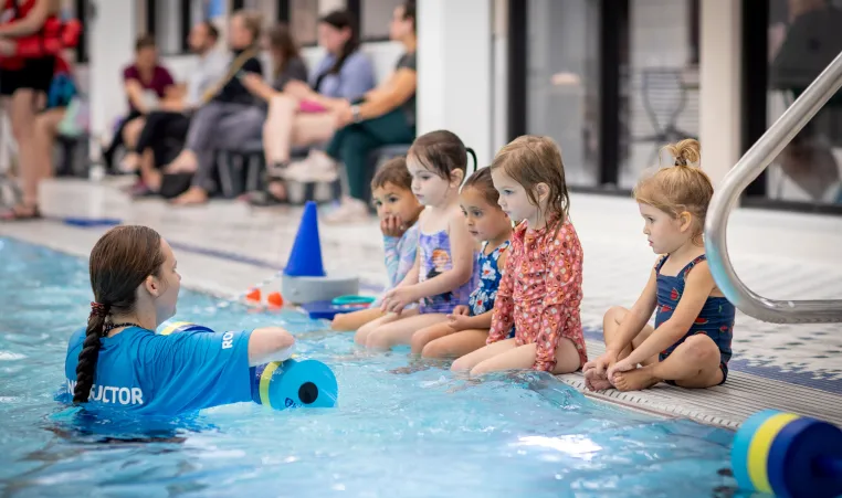 a swim instructor is in the pool speaking with a group of five young swimmers during a swim lesson