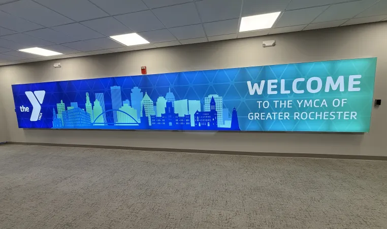 image of sign that reads "welcome to the YMCA of Greater Rochester" in blue and green with the skyline of Rochester