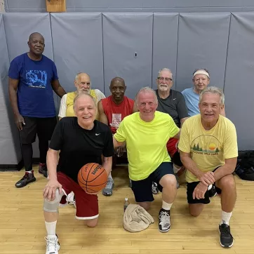 8 older men posing for a photo in the northwest gymnasium with a basketball