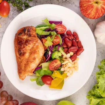 Plate full of healthy food items, including chicken, tomatoes, lettuce, peppers, and radish. It is surrounded by corn, forks, grapes, garlic, and lettuce on the surface holding the plate