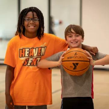 2 boys pose together smiling in the gymnasium, while the one on the right holds a basketball