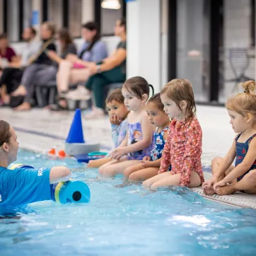 a swim instructor is in the pool speaking with a group of five young swimmers during a swim lesson