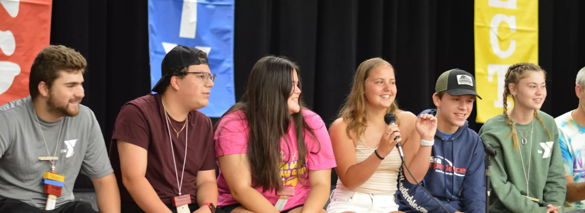 a group of teens sitting at the edge of a stage, one with a microphone