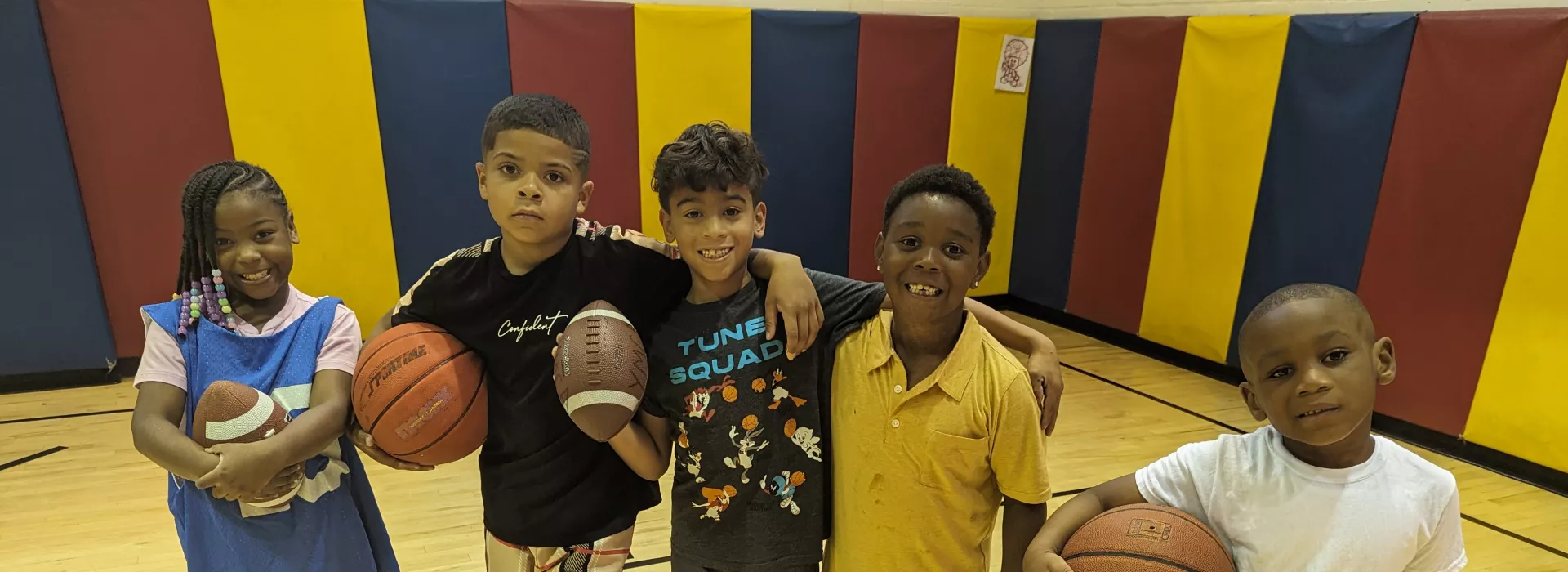 a group of 5 kids in the gym at the lewis street ymca neighborhood center. two kids are holding footballs, two kids are holding basketballs