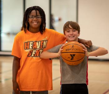 2 boys pose together smiling in the gymnasium, while the one on the right holds a basketball