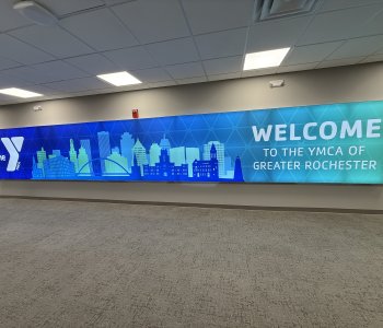 image of sign that reads "welcome to the YMCA of Greater Rochester" in blue and green with the skyline of Rochester